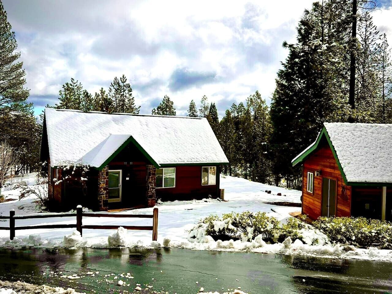 Two cabins with snow-covered roofs, a wooden fence, and a body of water in the foreground.