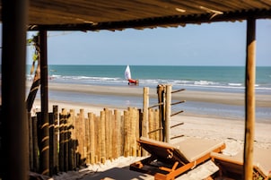 Vista a la playa con una cerca de madera, sillas de descanso y un velero en el agua.
