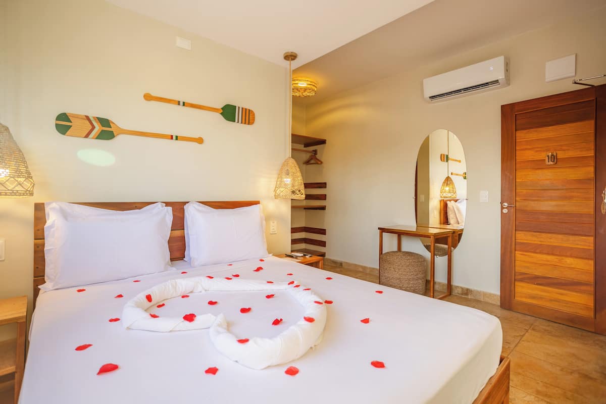 Una habitación de hotel con una cama grande, un cabecero de madera, una mesita de noche con lámpara, un espejo y una puerta de madera con un número.