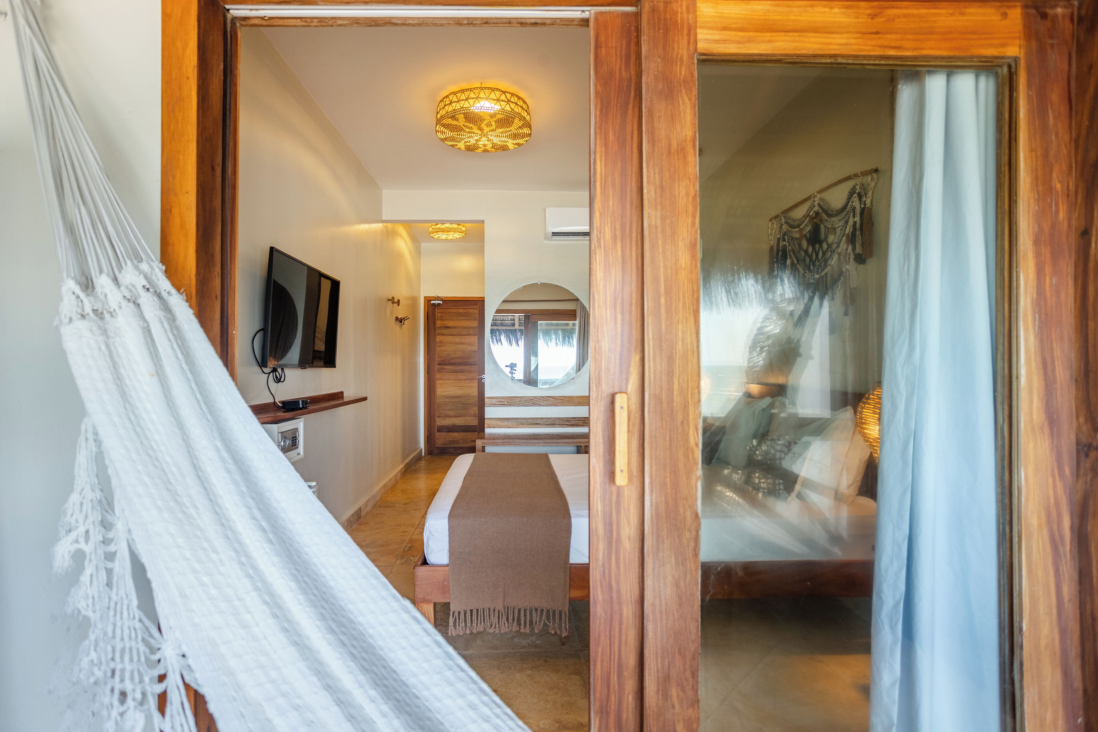 A room with a hammock, a bed with a brown blanket, a television on the wall, and a window with curtains.
