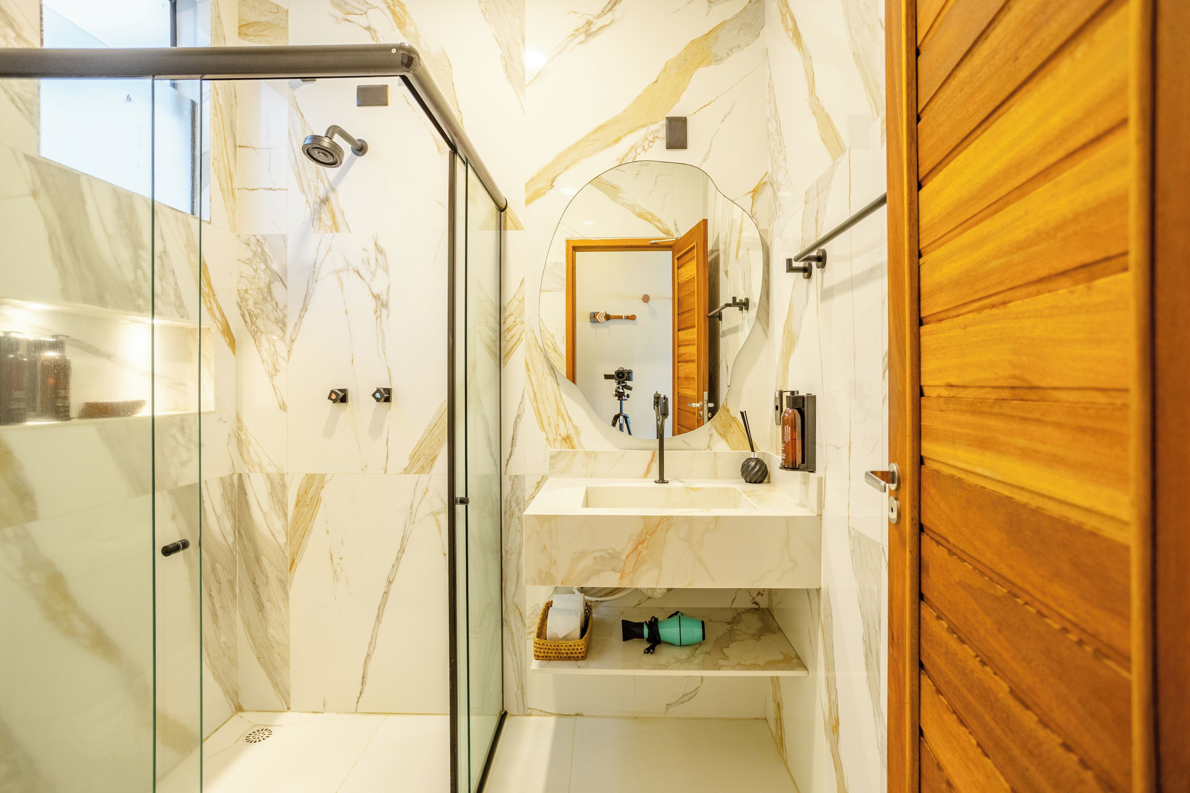 Un baño con ducha envuelta en cristal, pared de mármol, puerta de madera y espejo sobre el lavamanos.