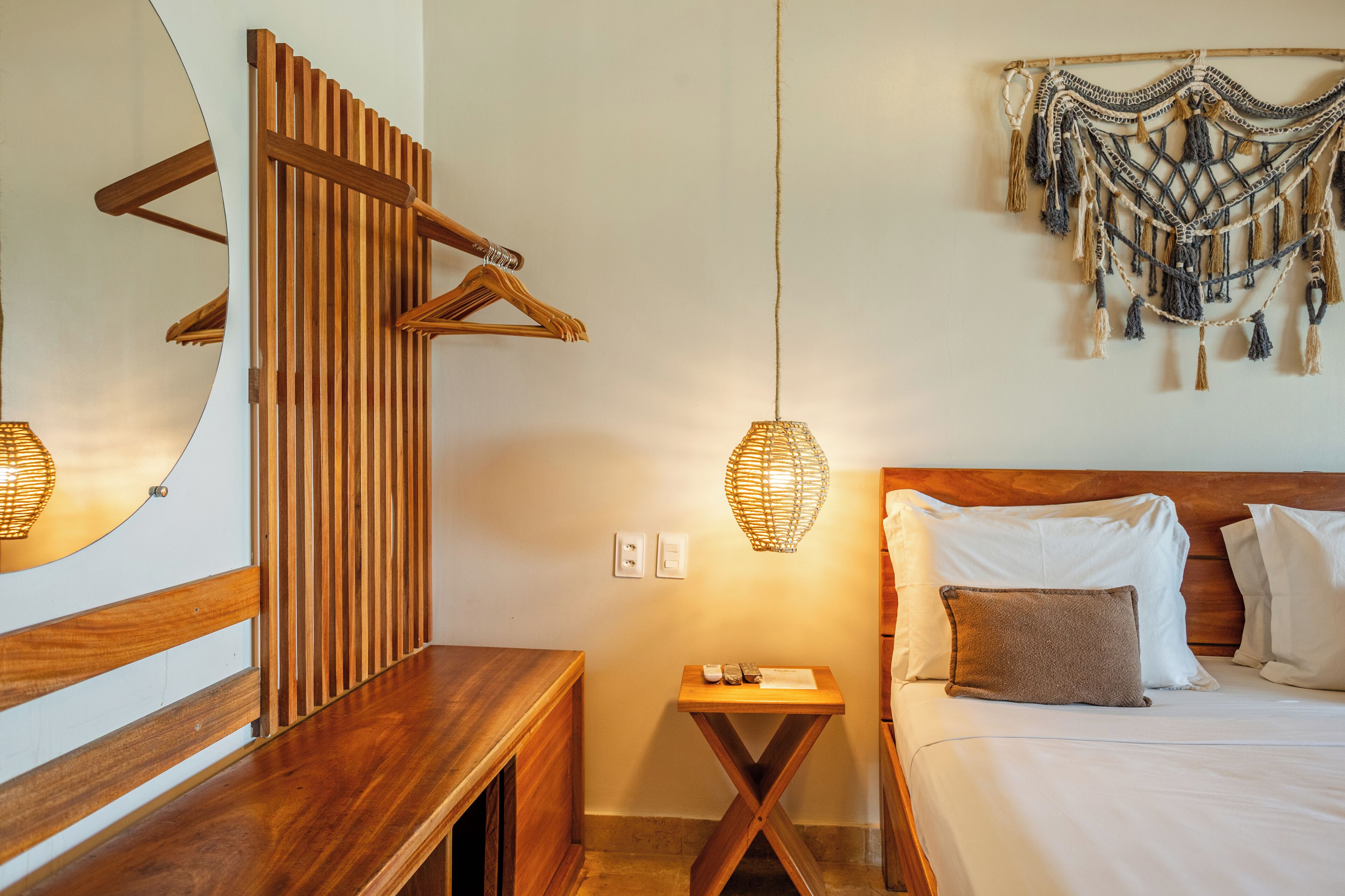 A neatly arranged bedroom with a wooden bench, a bedside table, a hanging light fixture, and a wall-mounted macrame piece.