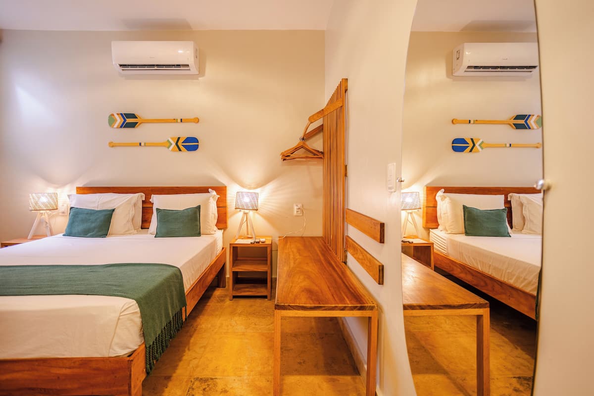 Habitación de hotel con dos camas, un banco de madera, una mesita pequeña y dos aires acondicionados instalados en la pared.