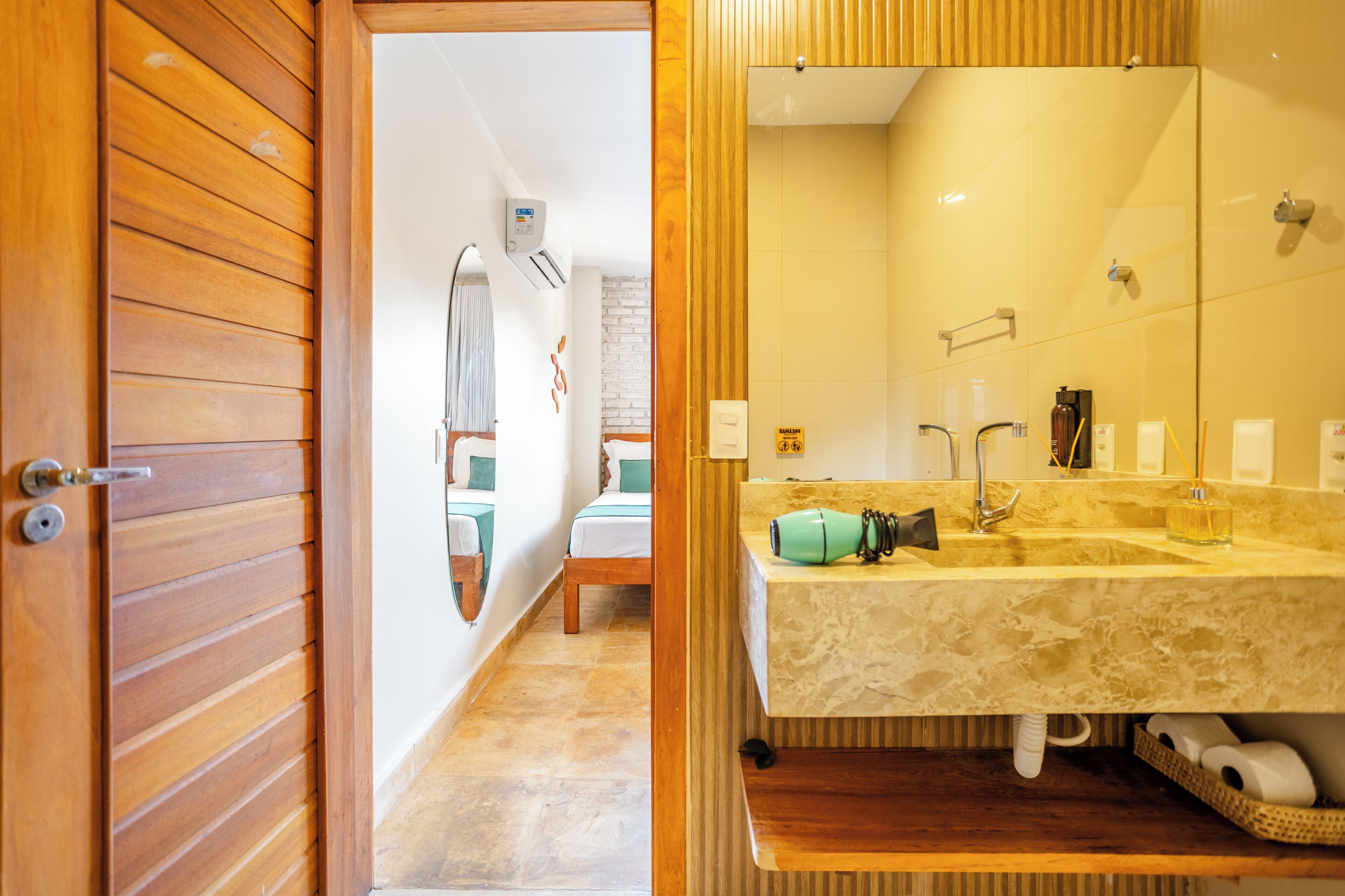 Une porte en bois donne sur une salle de bain équipée d’un grand miroir, d’un lavabo avec un distributeur de serviettes et d’un espace douche.