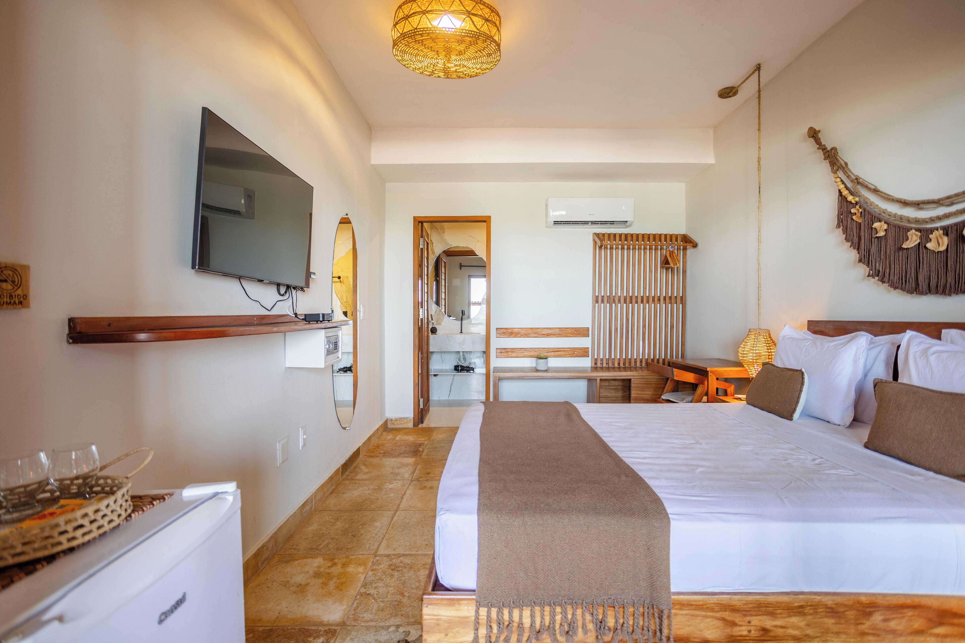 Quarto de hotel com uma cama grande, televisão na parede, um banco de madeira, uma mesinha e um ornamento decorativo.