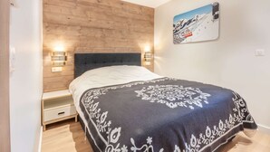 A bedroom with a wooden headboard, a bed with a patterned blanket, bedside lamps, and a wall-mounted picture of a ski lift. - Lapia104 : Splendid one bedroom + cabin (MORZINE)
