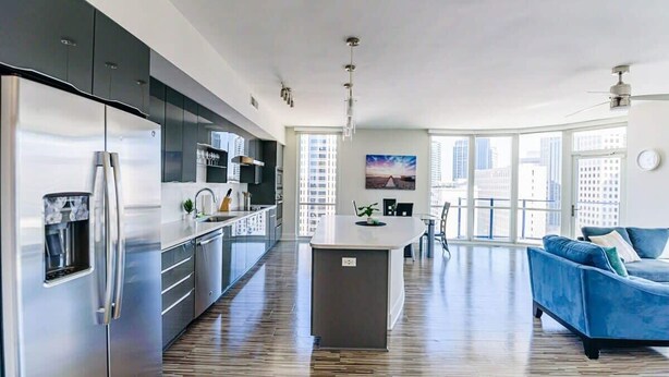 A modern kitchen with a large island, stainless steel appliances, and a blue sofa in a spacious living area with large windows. - Charlotte Uptown Fully Furnished Apartments (Charlotte)