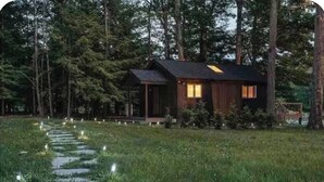 Une cabane aux murs en bois, entourée d’une forêt. Un sentier pavé de pierres mène à la cabane.