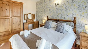 A neatly made bed with white linens and decorative pillows, a wooden dresser with a mirror, and a floral wallpaper. - Juice Trump - UK36670 (Belchford)