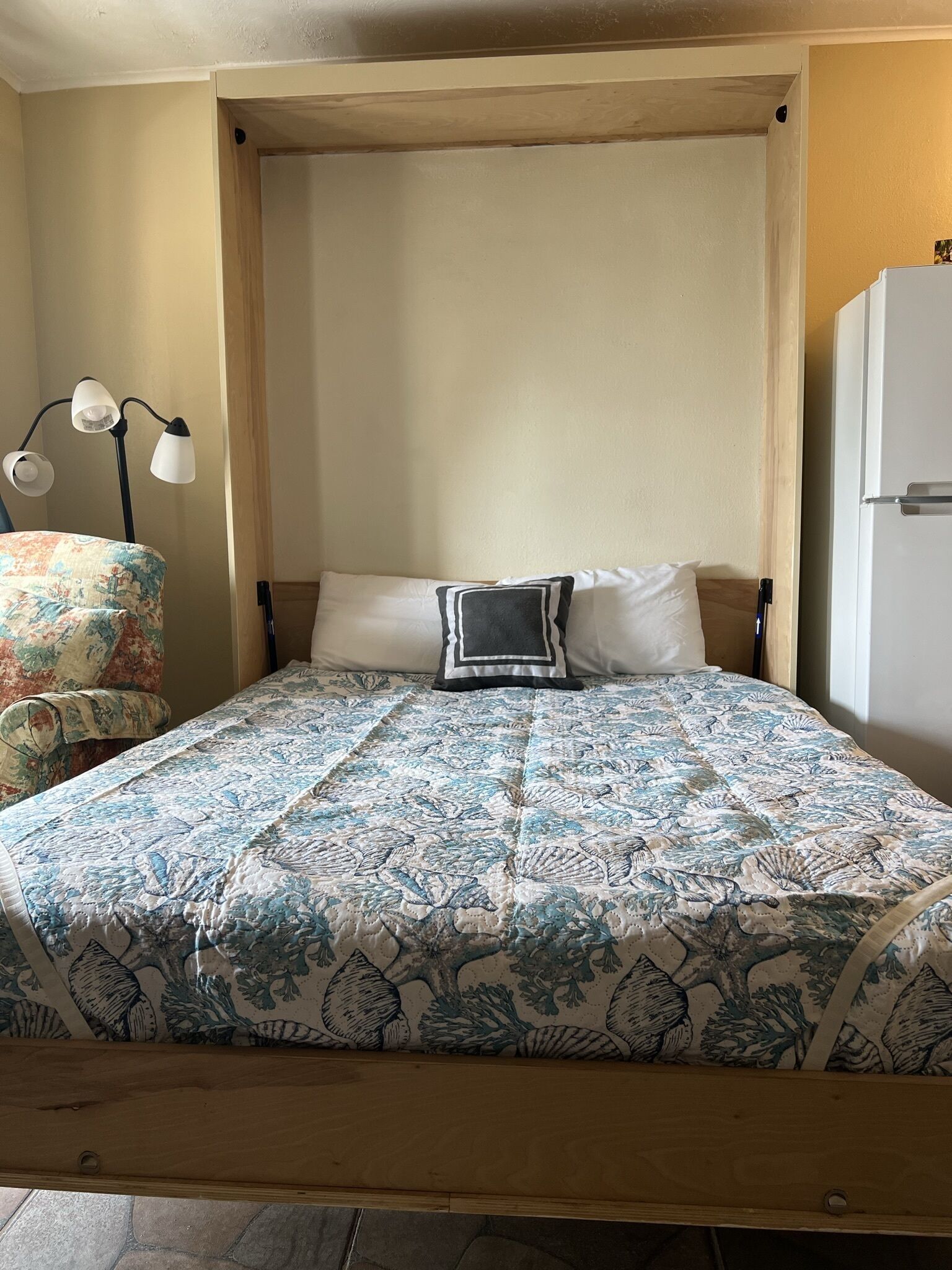 Een bed met een quilt met bloemenprint, een bedlamp en een stoel met een bloemkussen.
