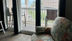 A room with a sofa, a chair, and a table near a sliding glass door leading to an outdoor balcony with a view of buildings and a body of water. - Lovely, beach view condo (Corpus Christi)