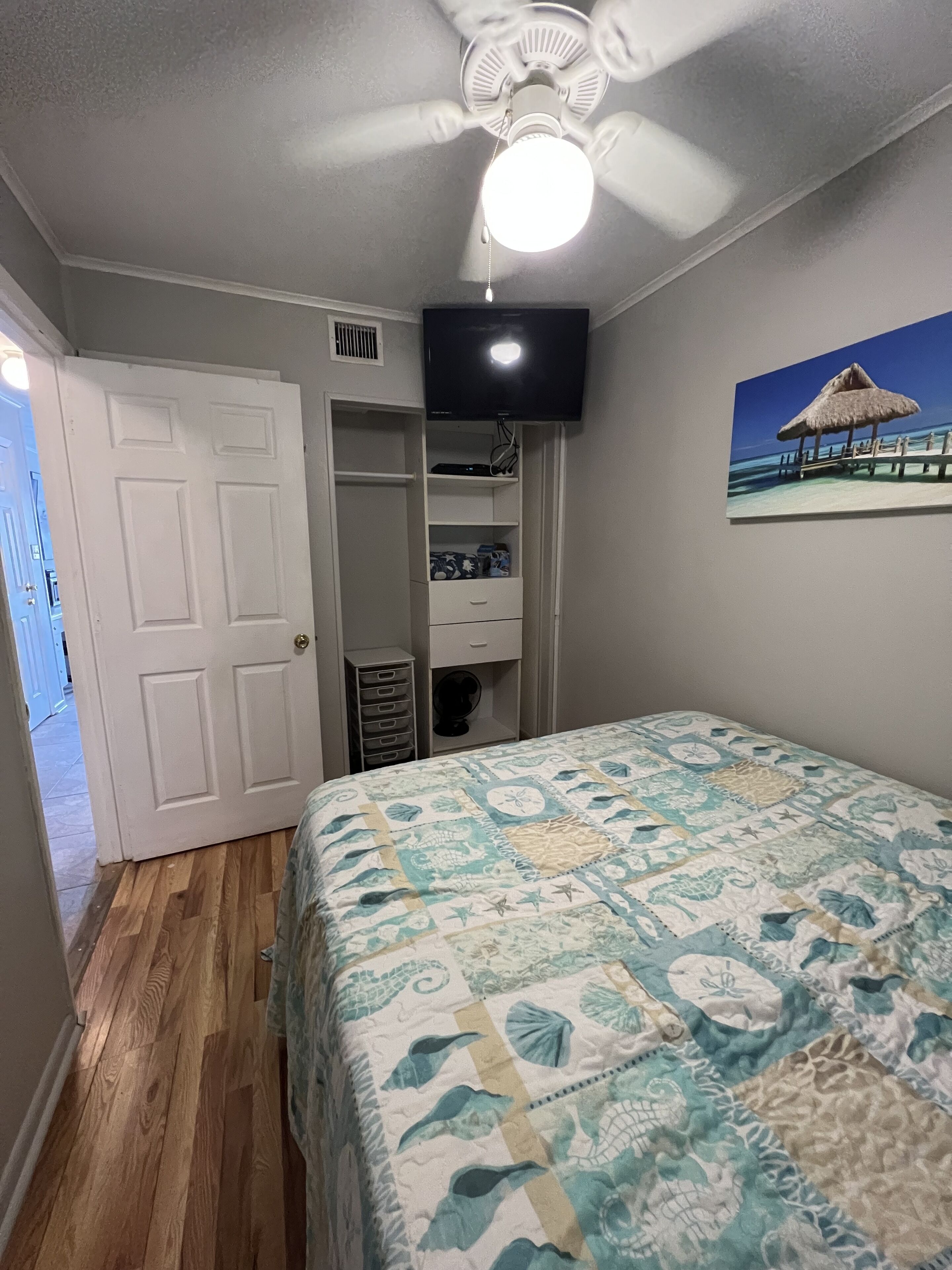 A bedroom with a bed, a ceiling fan, a TV mounted on the wall, and a painting of a beach scene.