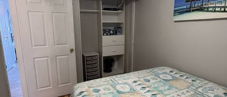 A bedroom with a bed, a ceiling fan, a TV mounted on the wall, and a painting of a beach scene.