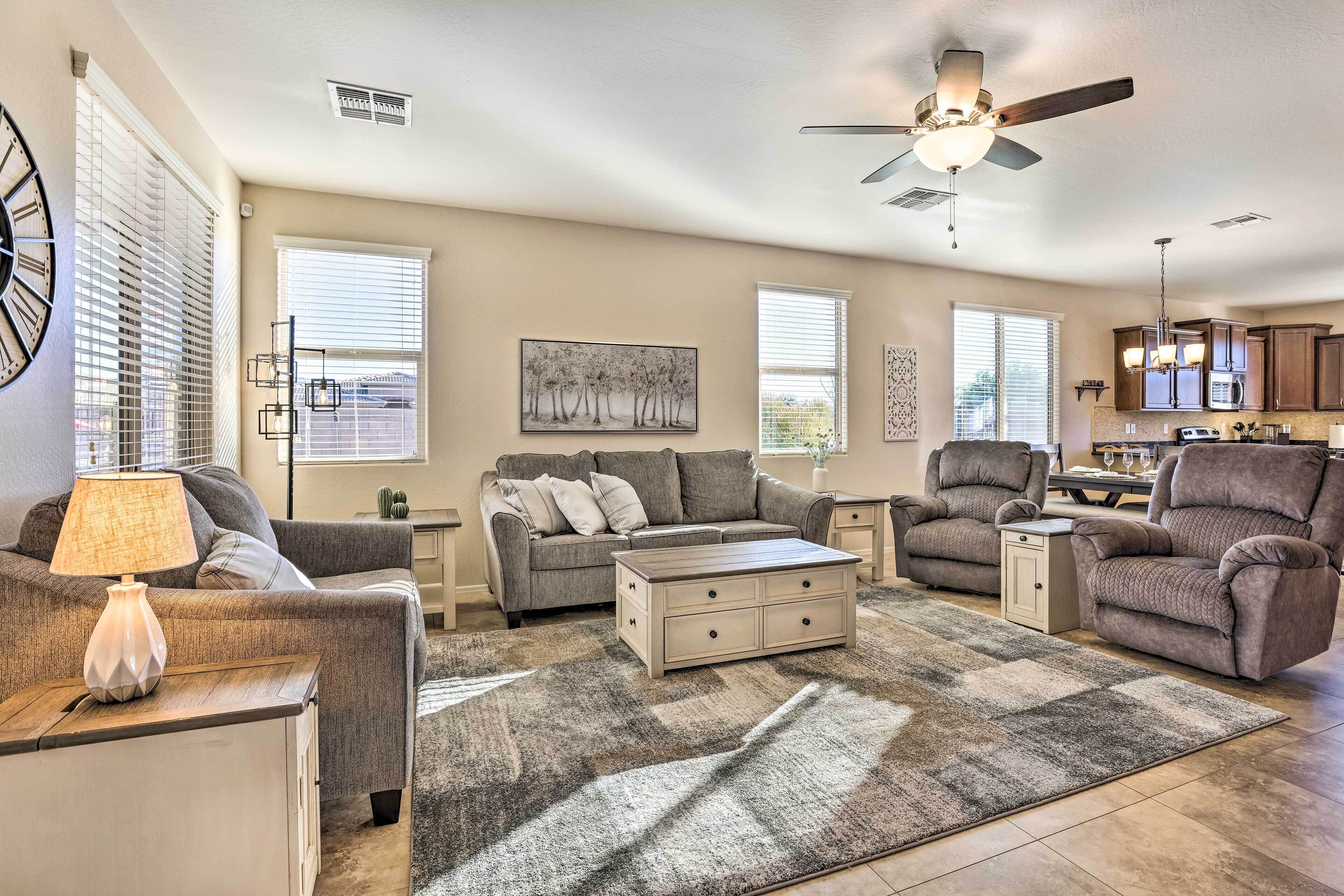 A living room with a sofa, armchairs, a coffee table, and a rug. There is a ceiling fan, a painting on the wall, and a kitchen area in the background.