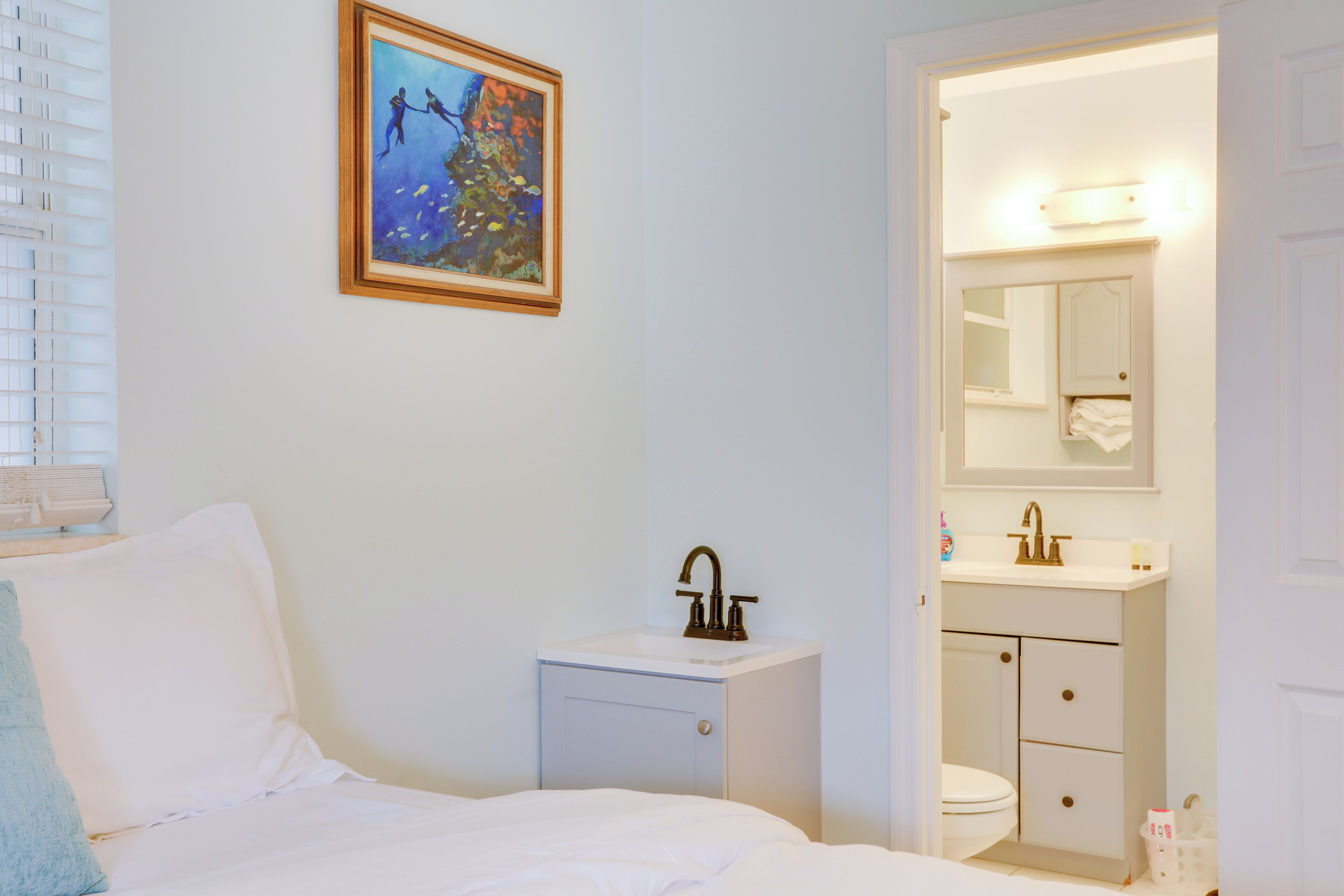Un dormitorio con cama, mesita de noche, un cuadro y baño con lavabo y espejo.