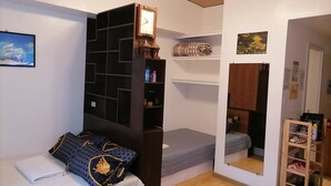 Quarto compacto com uma cama, estante embutida, um pequeno espaço de estar e um relógio fixado na parede.
