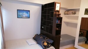Quarto compacto com uma cama, uma pequena estante, um relógio fixado na parede e um quadro emoldurado.