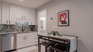 A modern kitchen with white cabinets, a herringbone tile backsplash, a stainless steel dishwasher, and a small dining area with a bench and a potted plant.