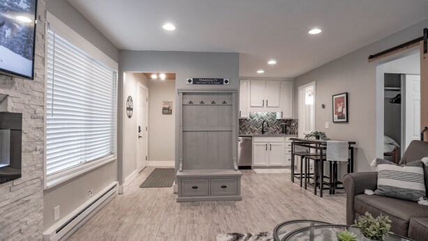 A modern kitchen with white cabinets, a built-in oven, and a herringbone backsplash. There is a dining area with a bar stool and a gray sofa.