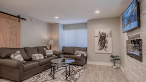 A living room with a brown sofa set, a glass coffee table, a patterned rug, a fireplace, and a TV mounted on the wall.