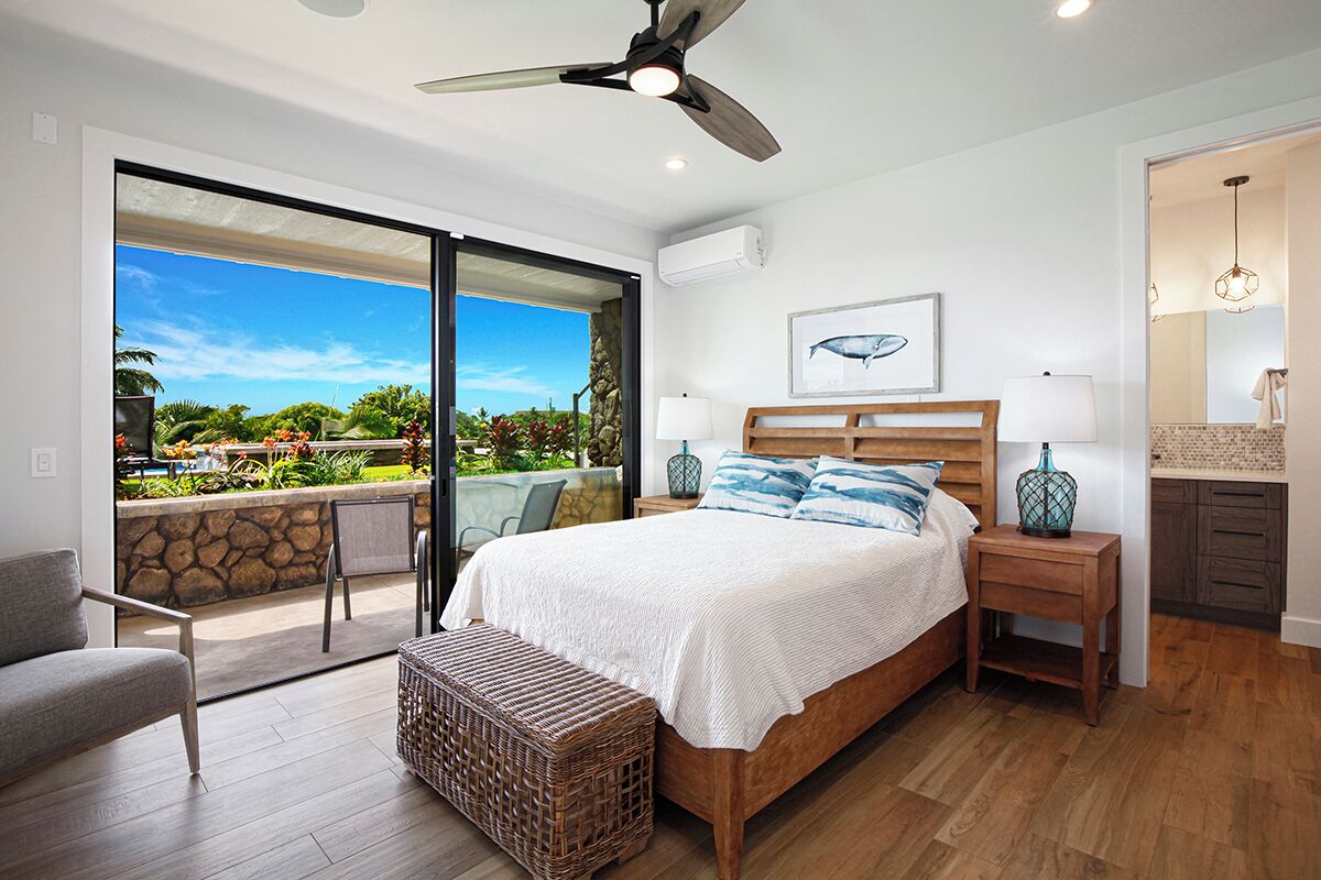 A bedroom with a large bed, a ceiling fan, a nightstand, and a view of a garden through a sliding door.