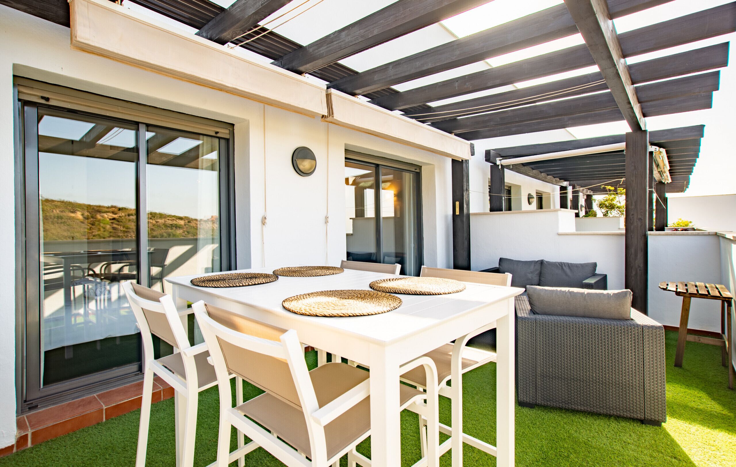 Eine Terrasse im Freien mit weißem Tisch und Stühlen, einer Korbus-Couch und einem Pergolen-Balkon.