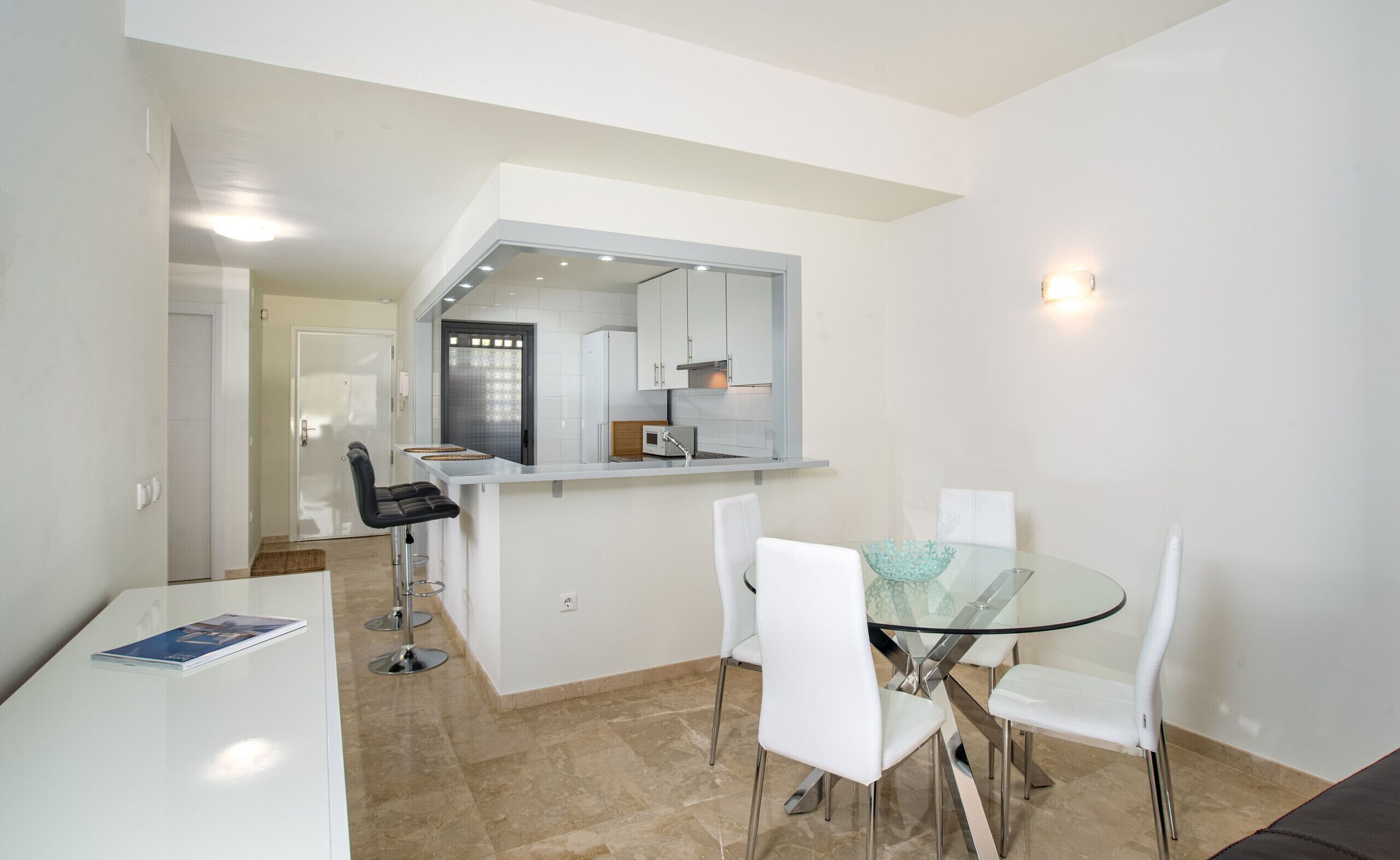 A modern kitchen with white cabinets, a built-in oven, and a microwave. There is a glass dining table with four white chairs and a bar area with a white countertop and stools.