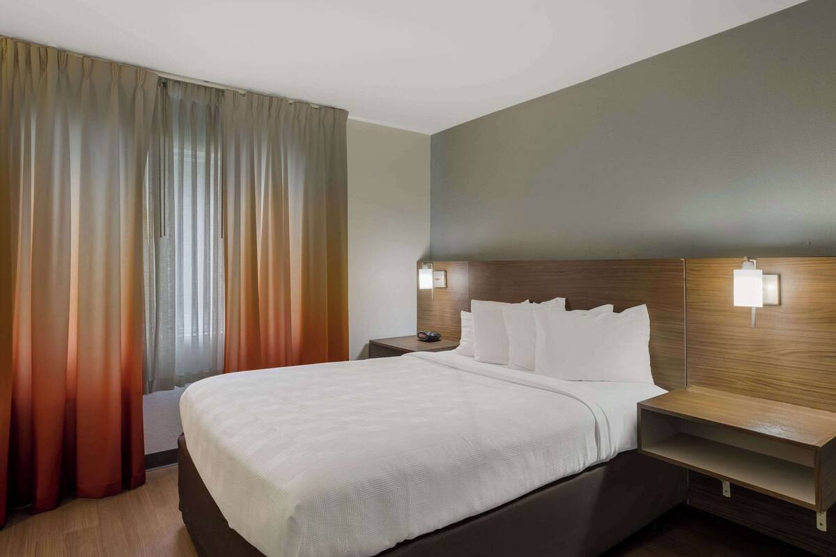 A hotel room with a large bed, two bedside tables with lamps, a wooden headboard, and a view of the outside through sheer curtains.