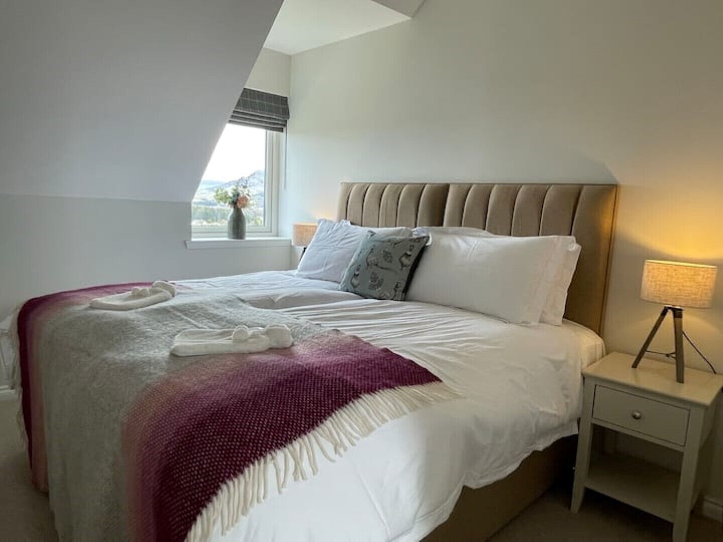 Una cama bien tendida con edredón blanco y manta morada, una lámpara de noche y una ventana con vistas al exterior.