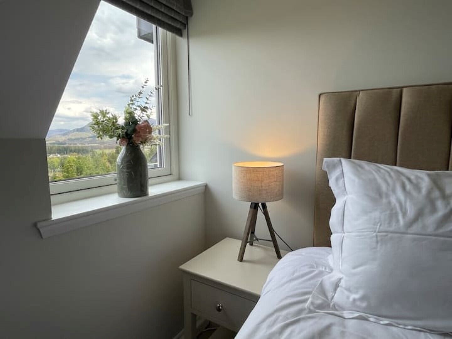 Ein Schlafzimmer mit Bett, Nachttisch mit Lampe, Fenster mit Blick auf Grünflächen und einer Blumenvase.