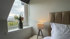 A bedroom with a bed, a bedside table with a lamp, a window with a view of greenery, and a vase with flowers.