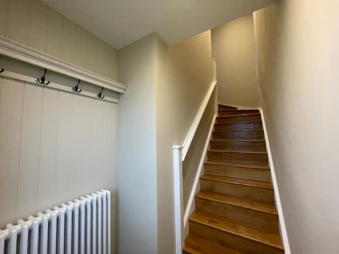 Eine Treppe mit Holzbodenstufen und weißen Geländern, die zu einer Zwischenebene mit einem weißen Heizkörper und Wandhaken führt.