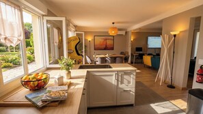 A modern kitchen with a dining area, a large window with a view of a garden, and a TV in the background.