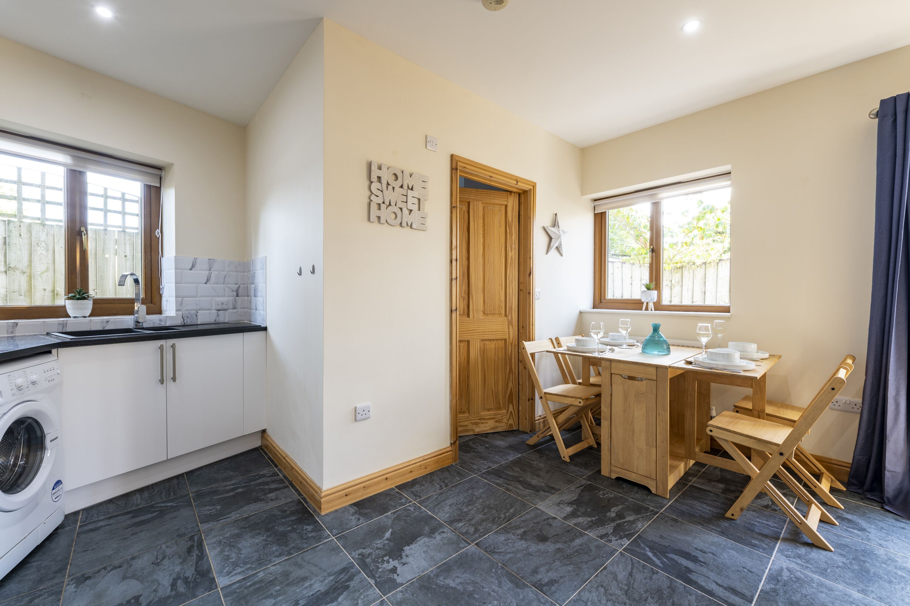 A compact kitchen with a washing machine, a dining area with a wooden table and chairs, and a wooden door.