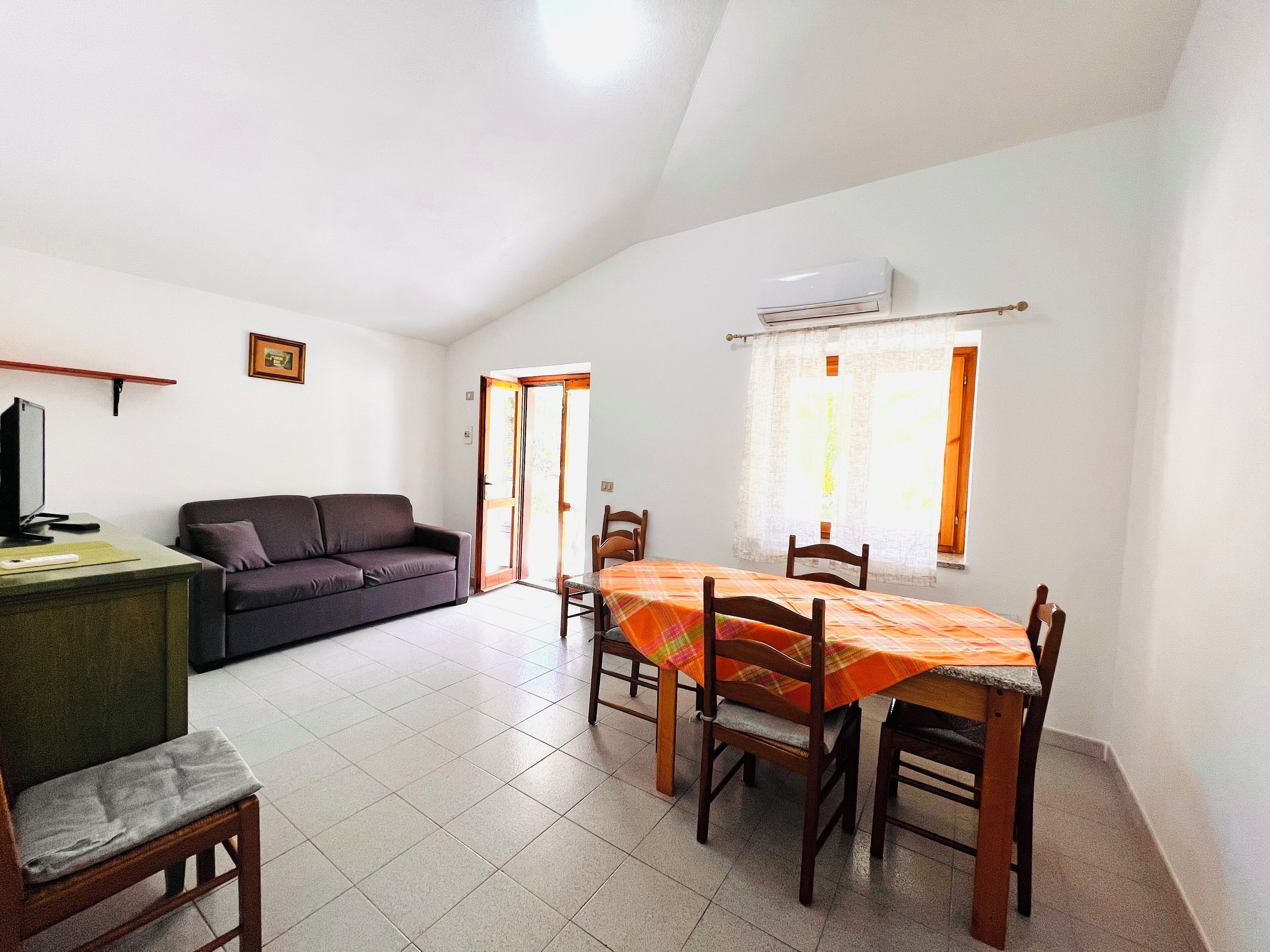 A spacious living area with a sofa, a dining table with chairs, a television, and a sliding glass door leading to an outdoor area.