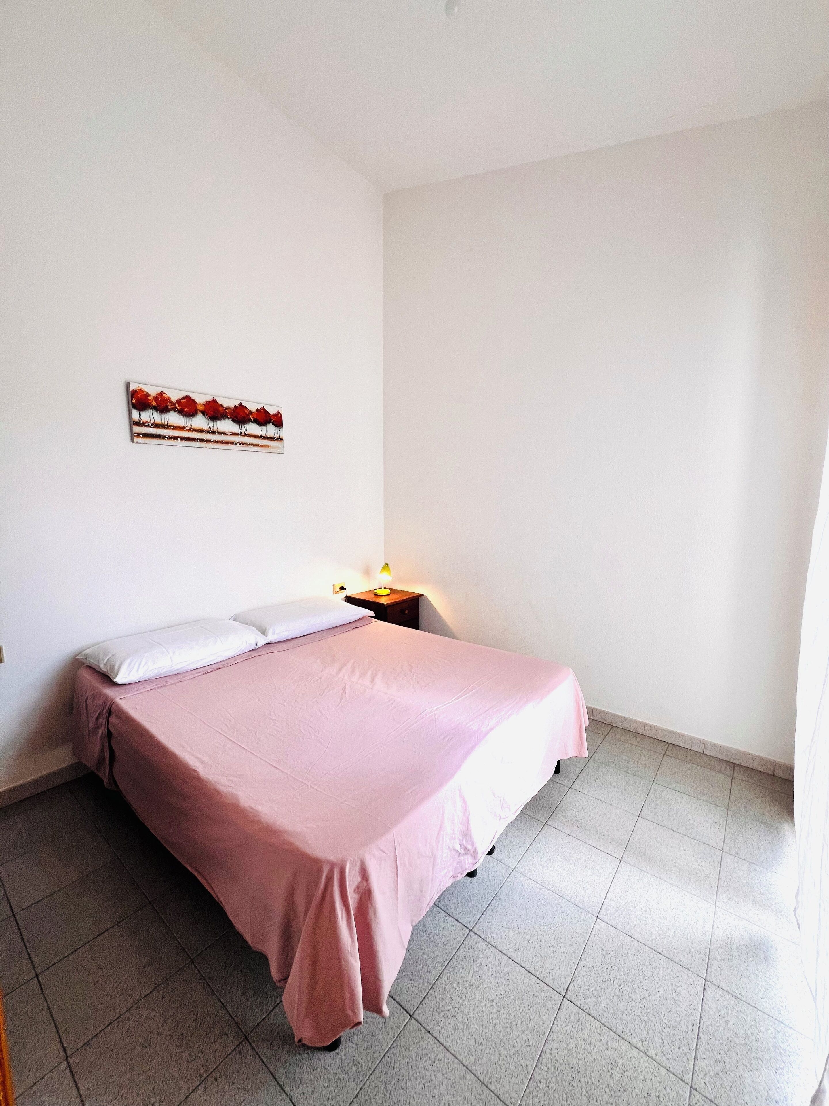 Un dormitorio con cama, mesita de noche, una obra de arte en la pared y suelo de baldosas.