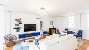 A modern living room with a white sofa, a TV, a bookshelf, and a dining area with chairs.