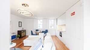 A modern living room with a white sofa, a dining area with blue chairs, a kitchen with white cabinets, and a ceiling light fixture.