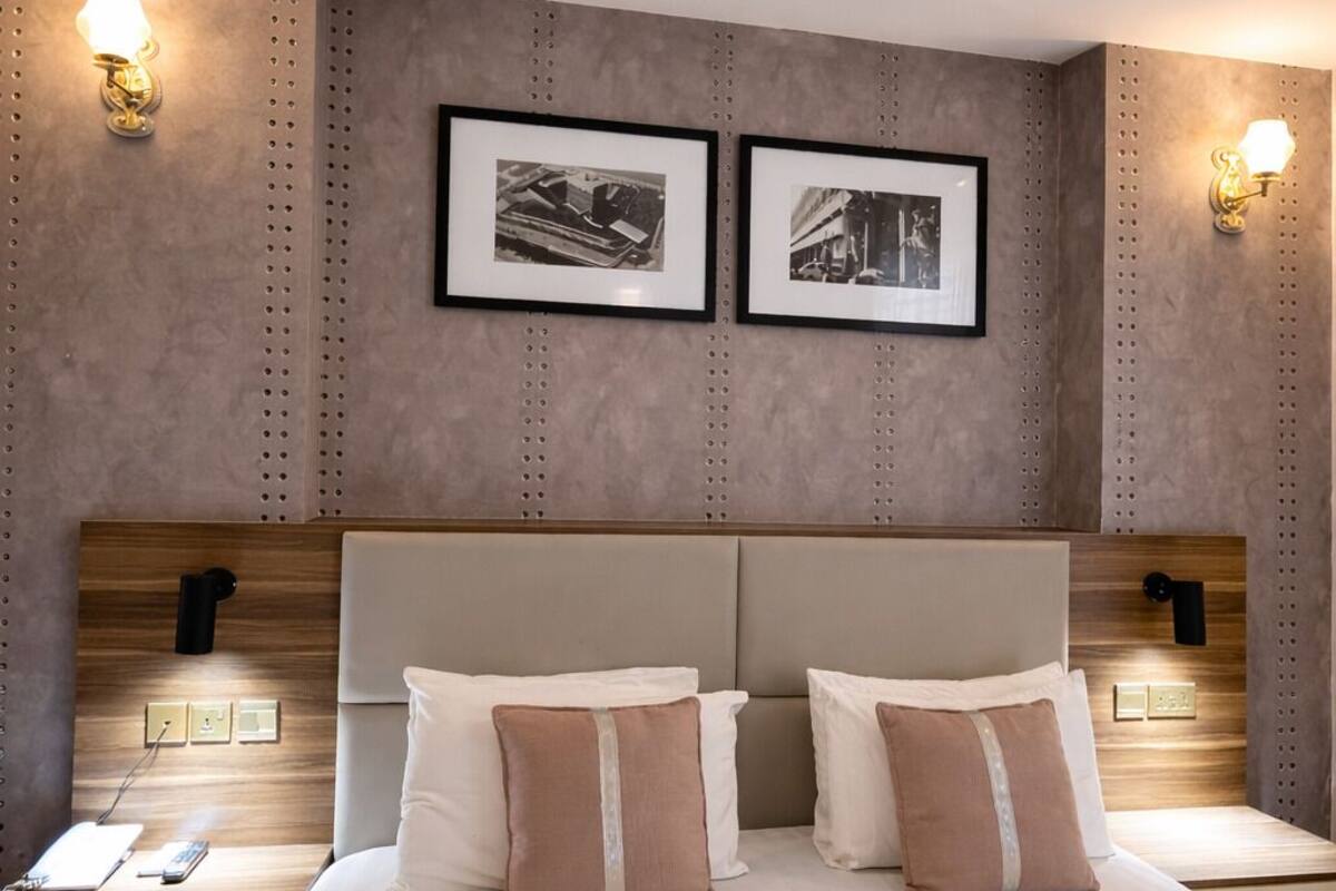 A hotel room with a large bed, two bedside tables, a headboard, wall-mounted lights, and two framed pictures.