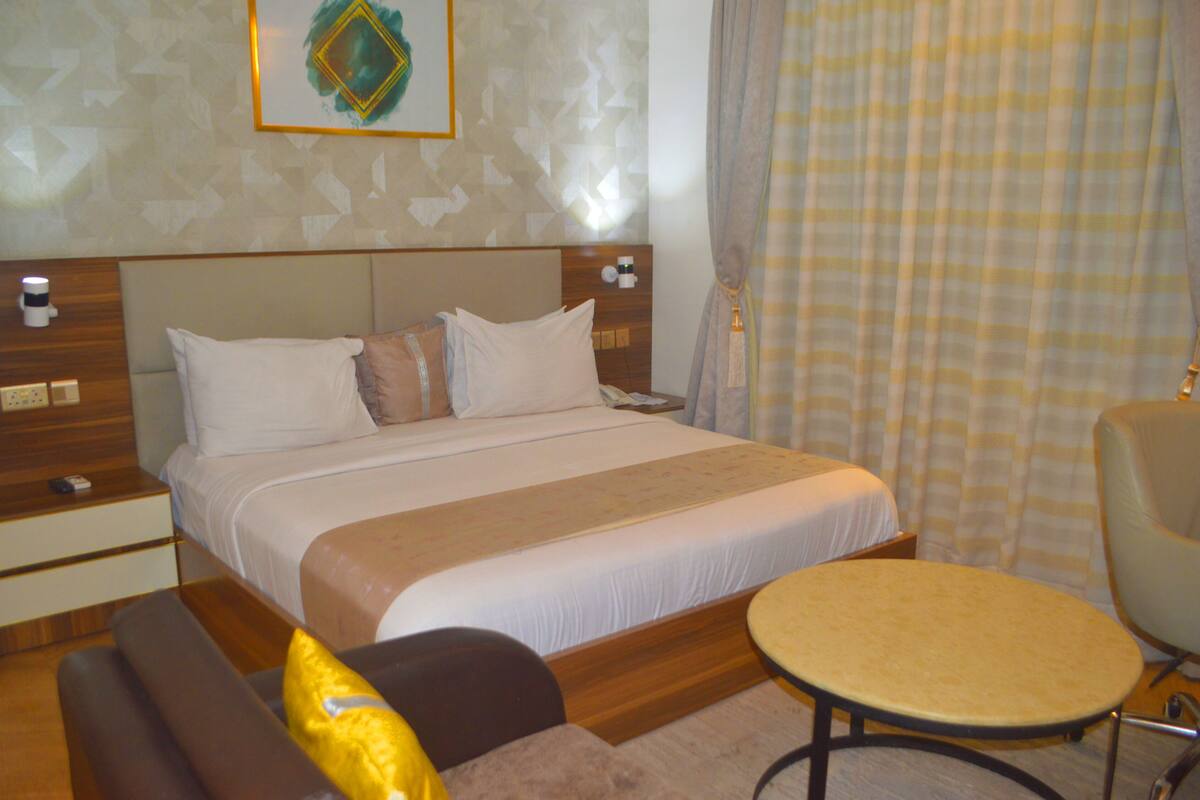A hotel room with a bed, bedside tables, a chair, a small table, and a curtain.