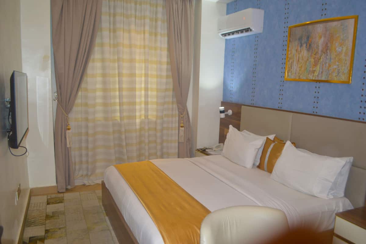 A hotel room with a bed, curtains, an air conditioner, a painting, and a telephone.