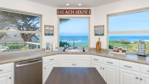 Una cocina con vista al océano, armarios blancos, una encimera de madera y un letrero que dice 'BEACH HOUSE'.