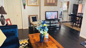 Una sala de estar con un sofá azul, una mesa de centro de madera con un florero, un televisor, una zona de comedor con sillas y una puerta.