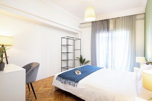 A neatly arranged hotel room with a bed, a desk, a chair, a plant, and a shelf.