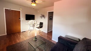 A living room with a glass coffee table, a black chair, a dark sofa, a ceiling fan, a mounted TV, and a wooden door. - Red Roof Rental (Thunder Bay)