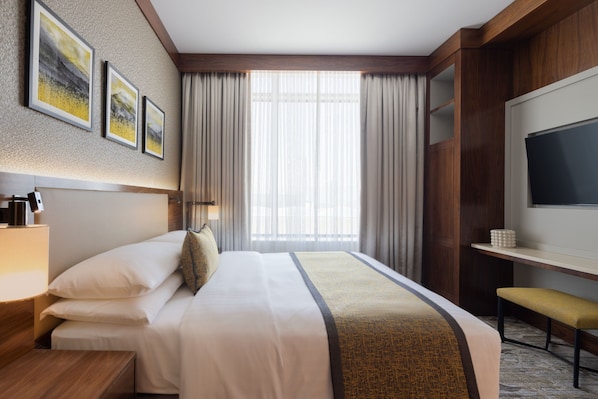 Habitación de hotel con una cama grande, un escritorio con silla, televisión y ventana con cortinas.