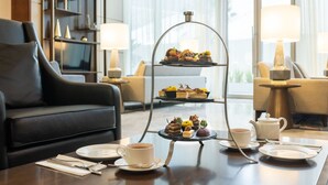 Ein dreistöckiges Kuchenbuffet mit verschiedenen Backwaren und einem Teekännchen auf einem Tisch in einem modernen Hotelzimmer mit einer Ledersofa und Lampen.