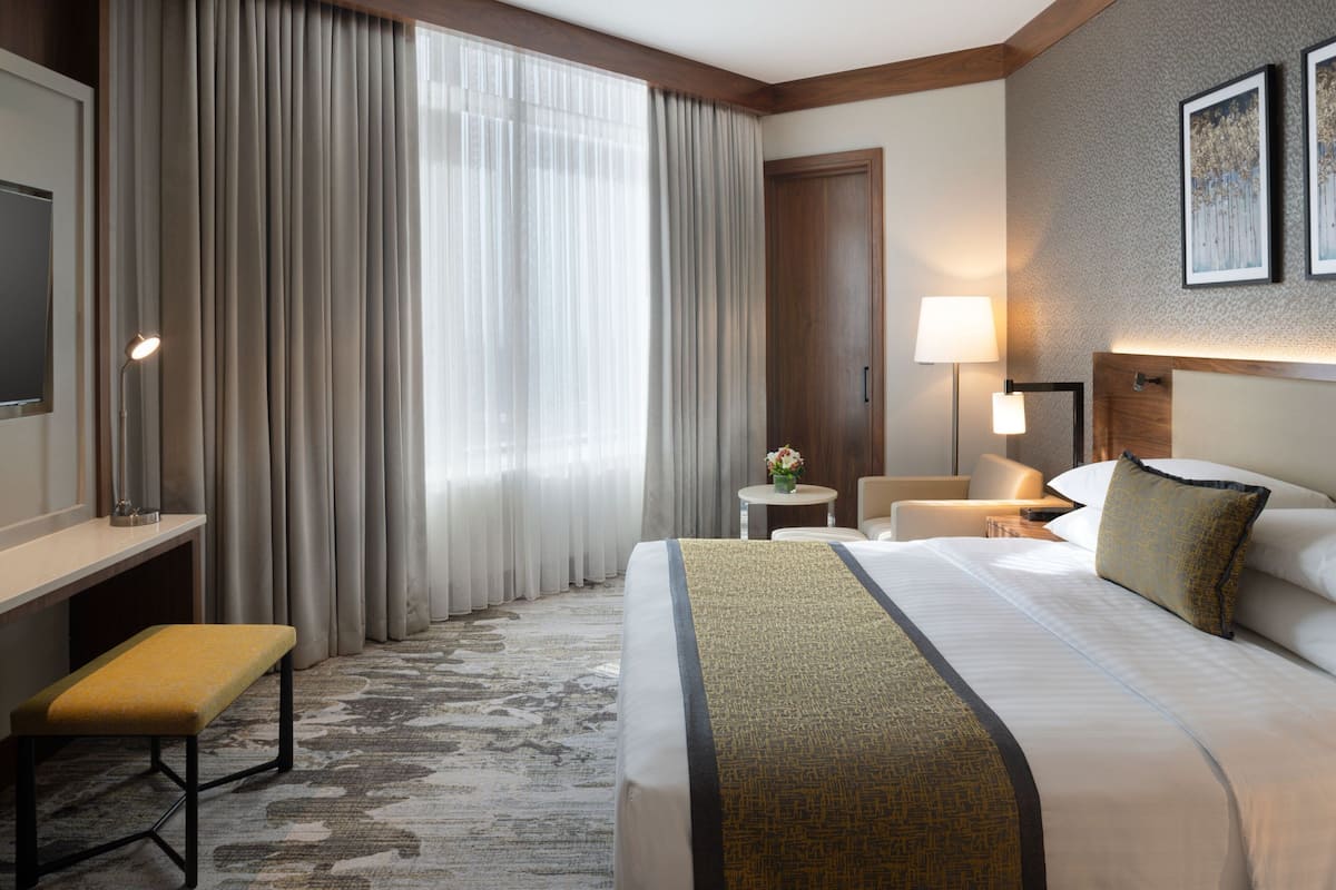 Ein Hotelzimmer mit einem großen Bett, einem Schreibtisch, einem Sessel, einer Lampe und einem kleinen Tisch mit Blumen.