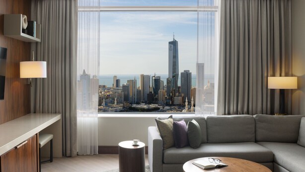 Ein modernes Hotelzimmer mit großem Fenster und Blick auf die Stadt, einer Couch, einem kleinen Tisch und einem Beistelltisch.