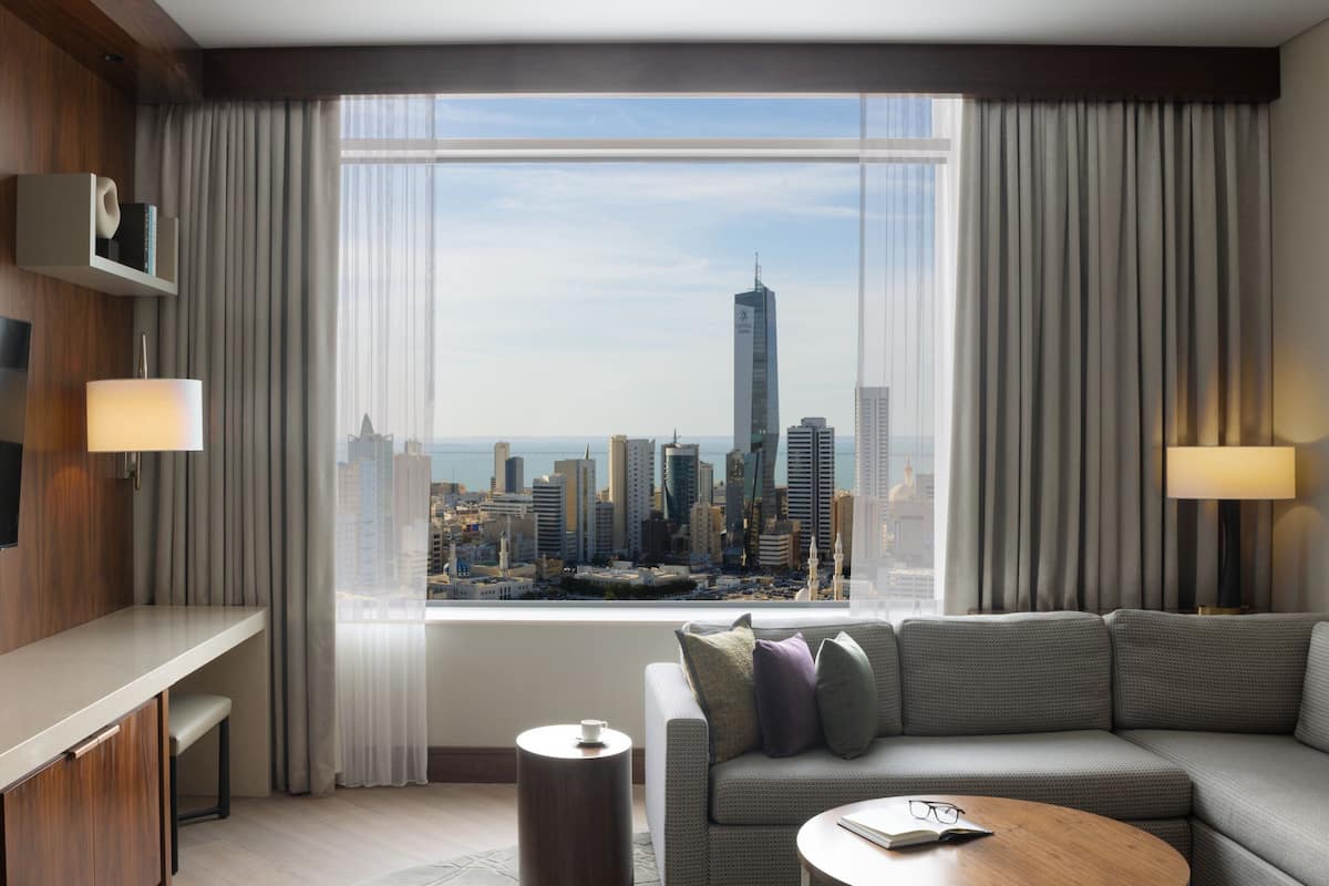 Ein modernes Hotelzimmer mit großem Fenster und Blick auf die Stadt, einer Couch, einem kleinen Tisch und einem Beistelltisch.